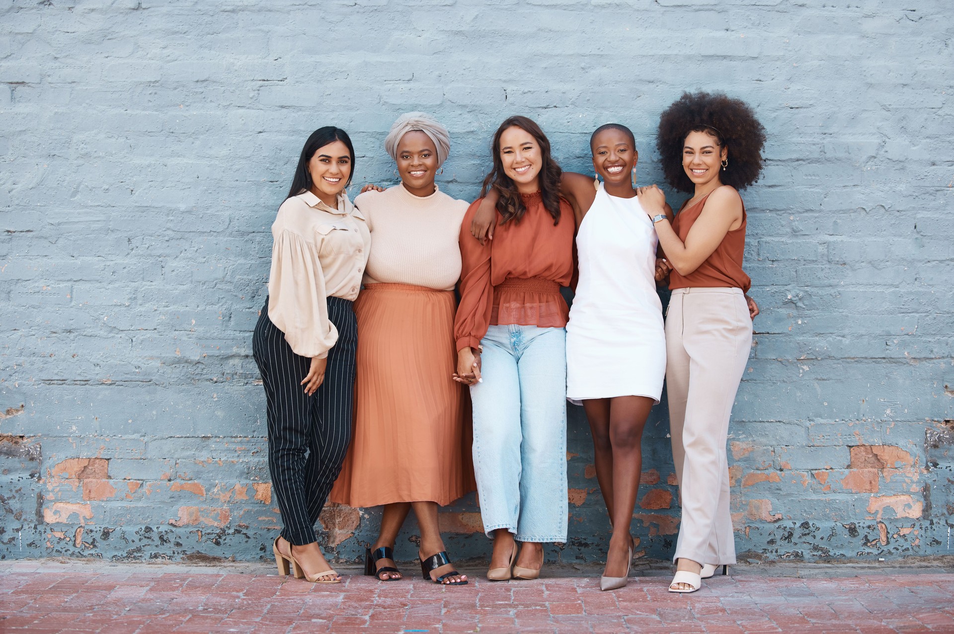 Group of five young happy cheerful businesswomen hugging while standing against a wall outside in the city. Portrait of happy colleagues holding hands and smiling standing in a line outdoors